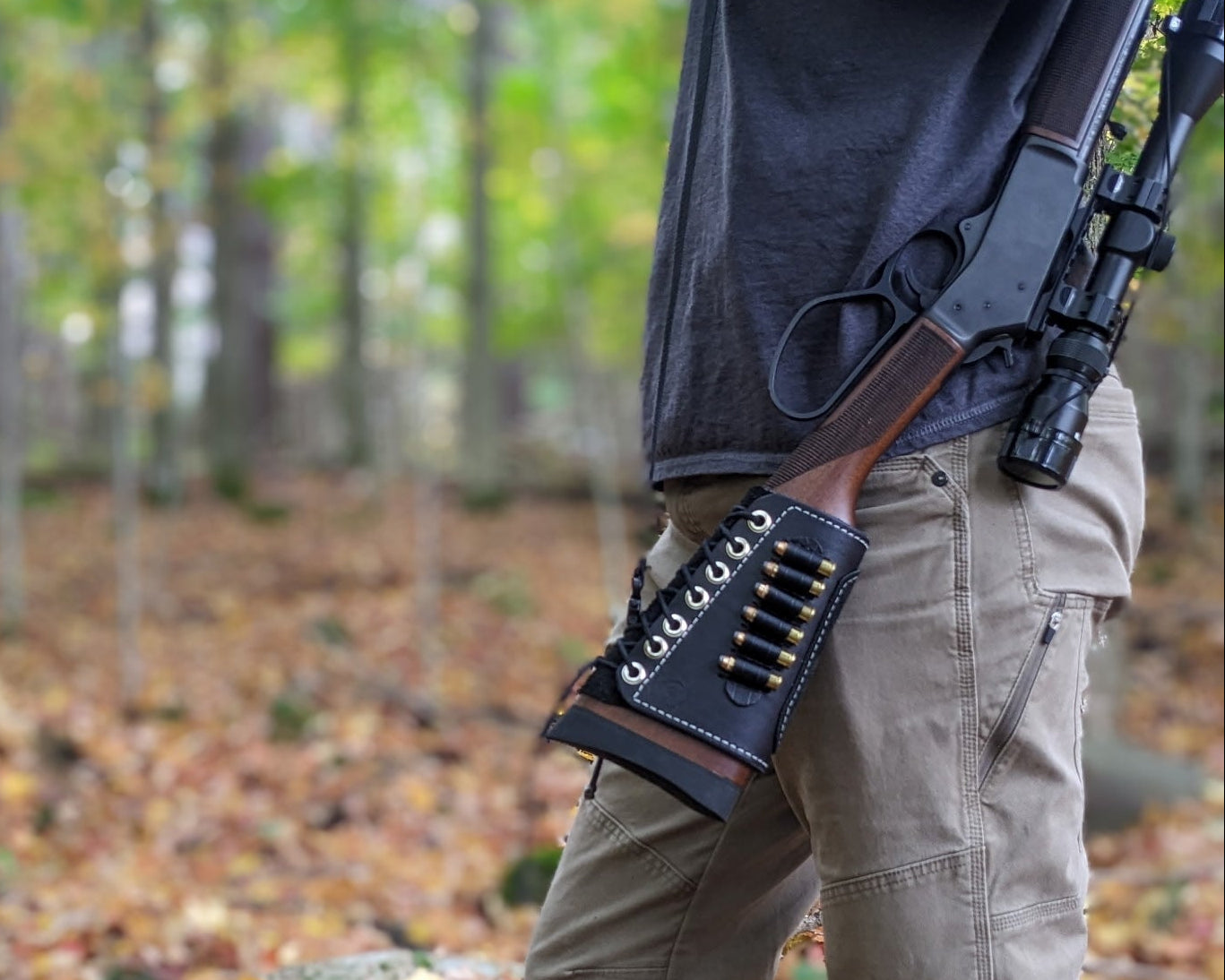 Person in a forest holding a rifle with a scope, standing next to a rock.