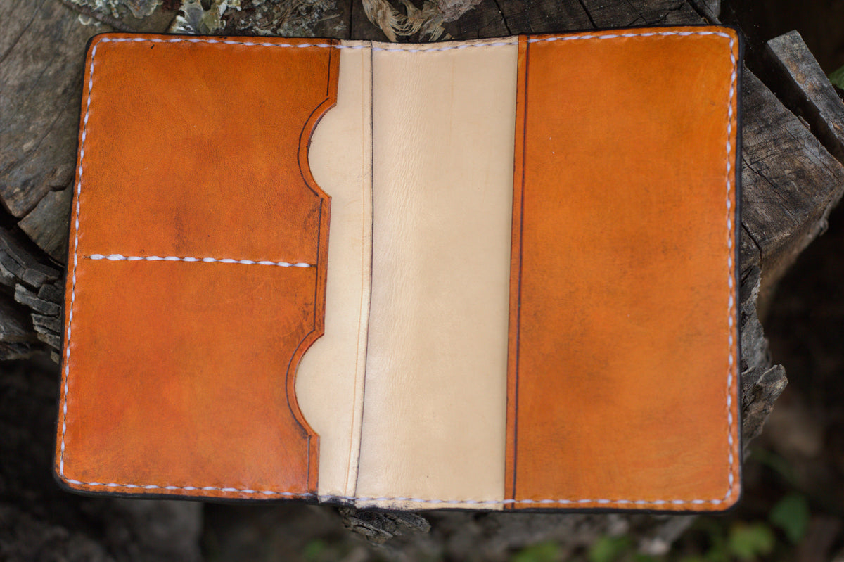 A handmade leather notebook cover or passport holder in natural tan and light beige, displayed open on a rustic tree stump.