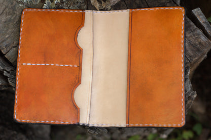 A handmade leather notebook cover or passport holder in natural tan and light beige, displayed open on a rustic tree stump.