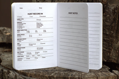 An open white hunting log notebook showing printed fields such as hunt record number, date, temperature, land type, game type, wind direction and speed, sky, moon phase, butch phase, and deer size, photographed standing on a rustic wood surface.